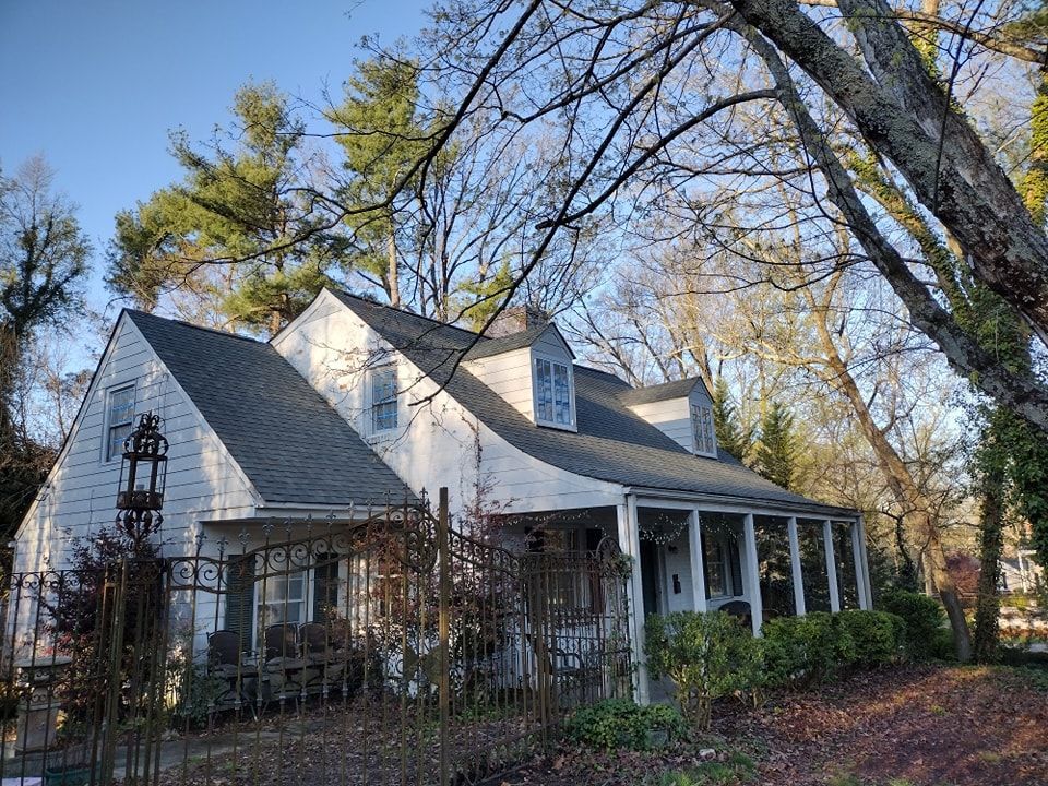 White house with dark roof and porch, surrounded by trees under a blue sky.