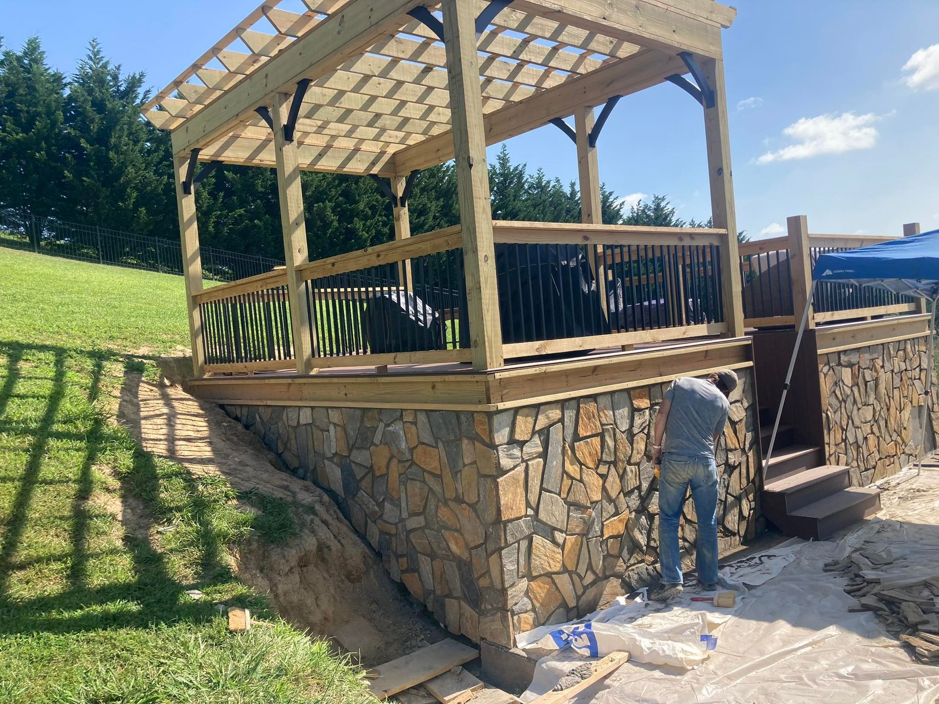 A man working on a stone wall, supporting a wooden deck with a pergola, set on a grassy hill.