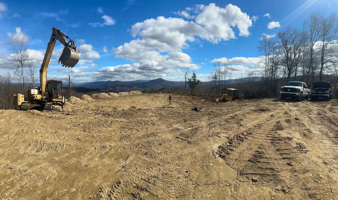 Construction site with excavator and truck under a cloudy blue sky. Brown dirt and clearing with mountain view.