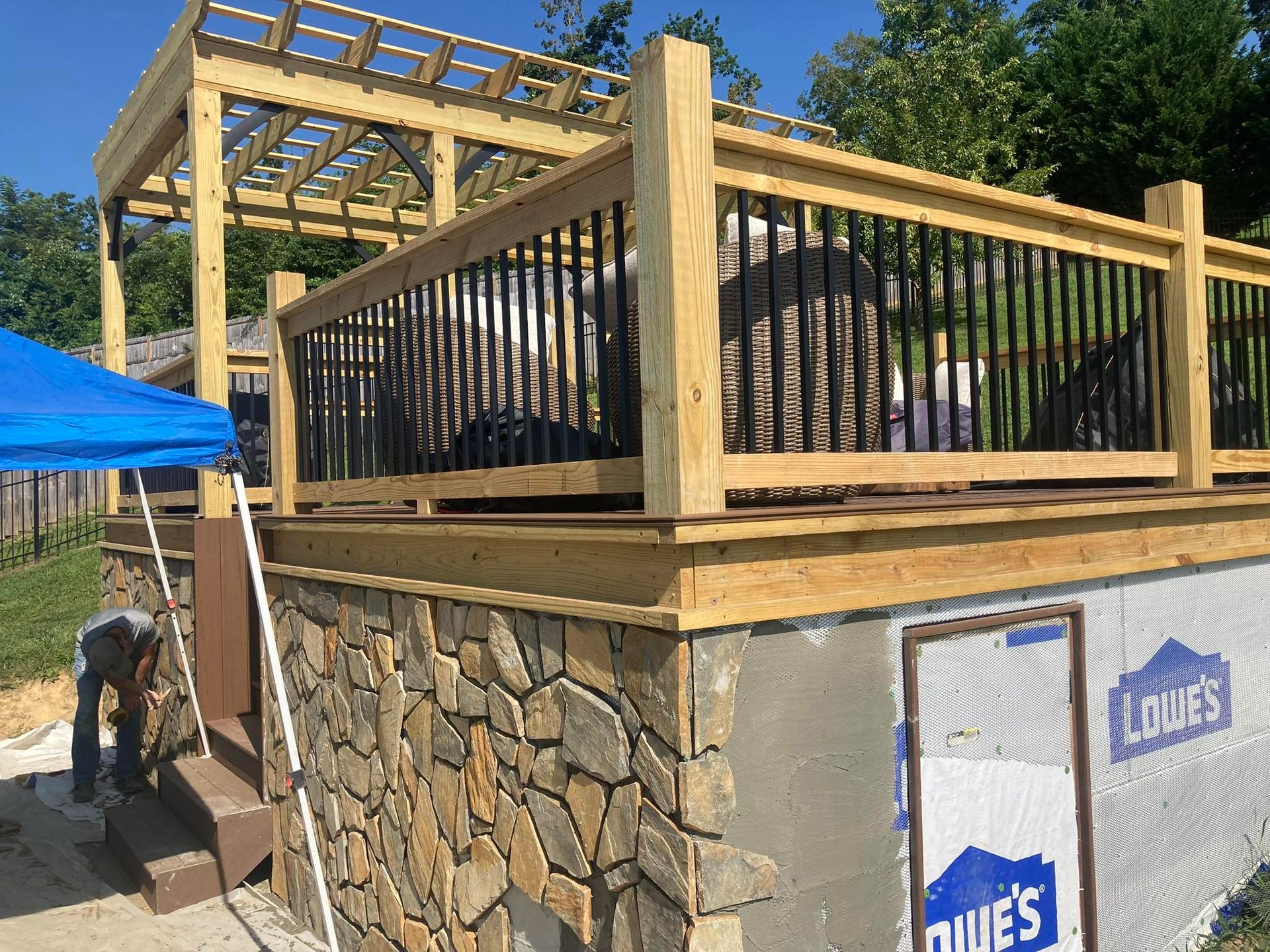Stone-faced deck with pergola, black railing, and a person working. Blue tarp in the foreground.