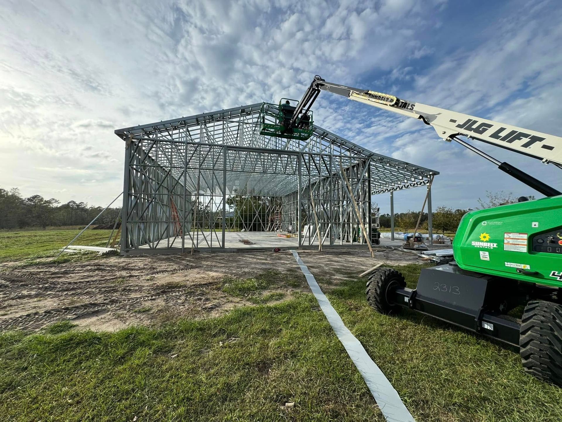 Metal-framed building under construction, workers on a lift, on a grassy field, cloudy sky.