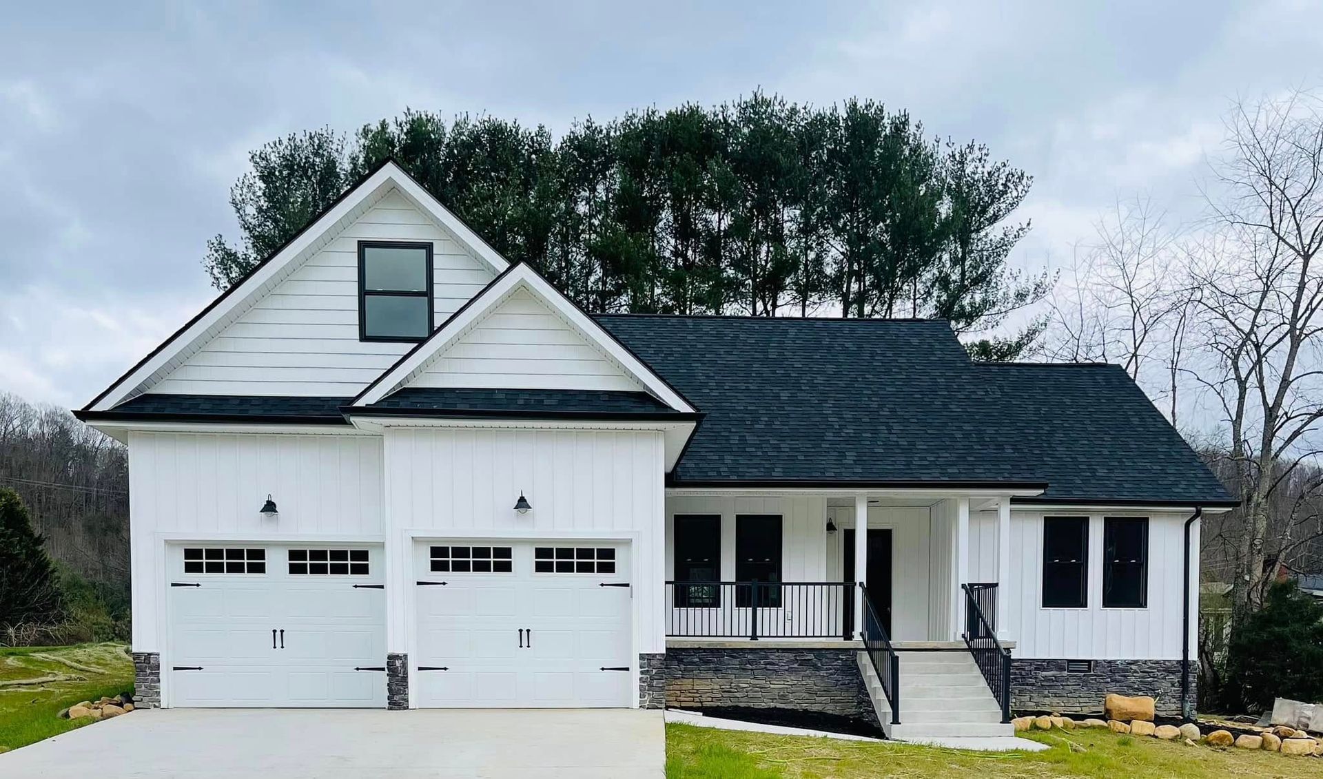 White farmhouse-style house with dark roof and trim, two-car garage, and stone accents under a cloudy sky.
