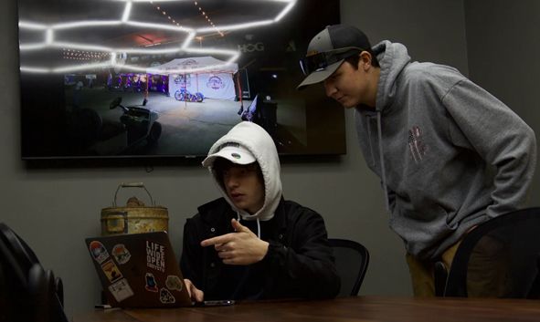 a man in a hoodie is pointing at a laptop while another man looks on 