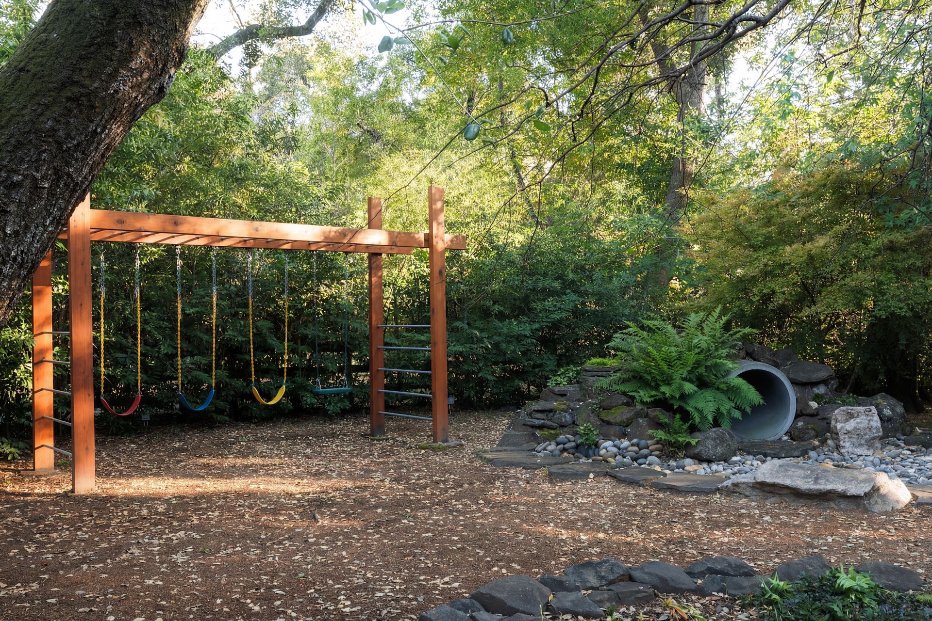 A wooden swing set in a wooded area with a small tunnel and lush foliage.