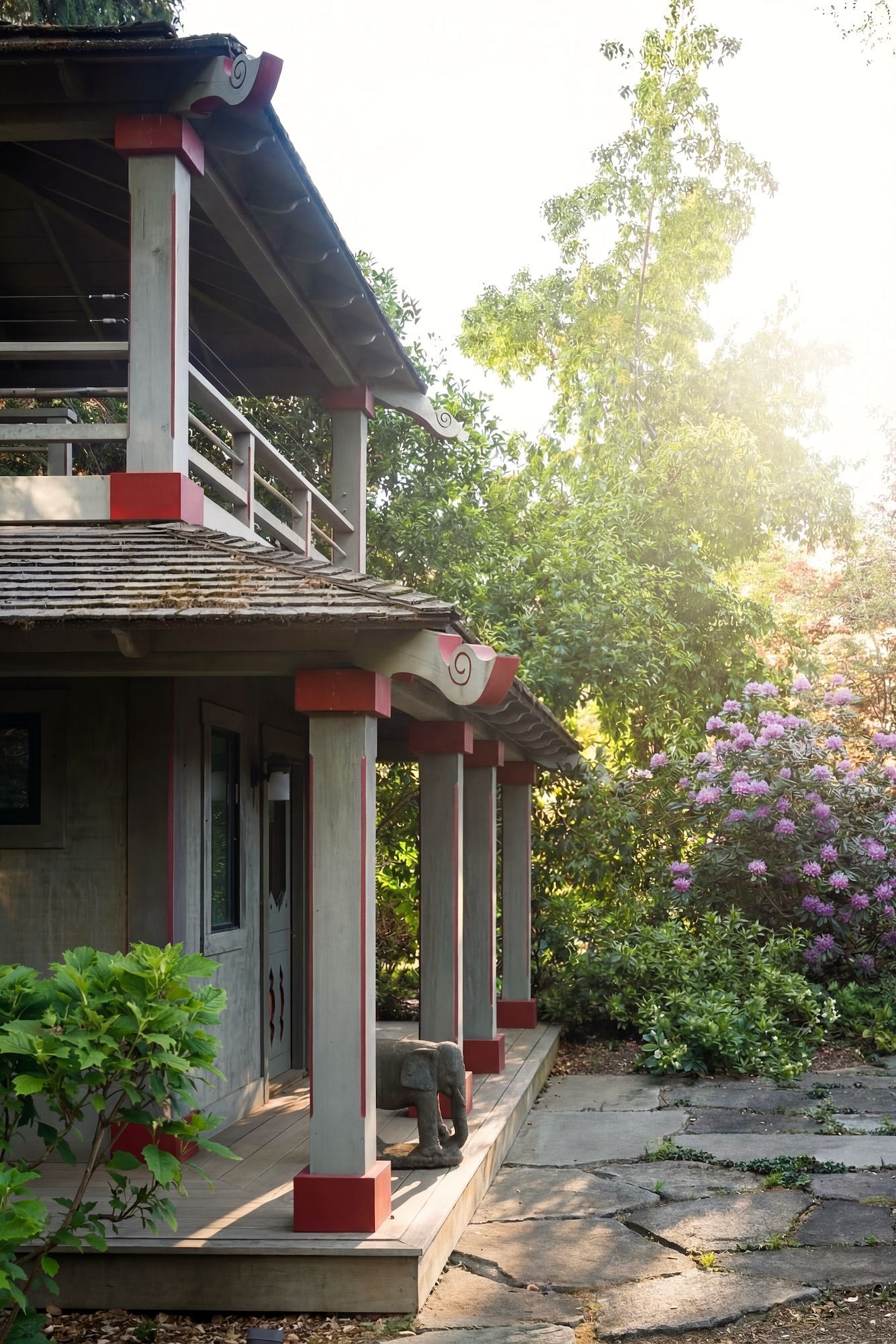 Asian-style building with red accents, porch, and elephant statue, surrounded by trees and sunlight.