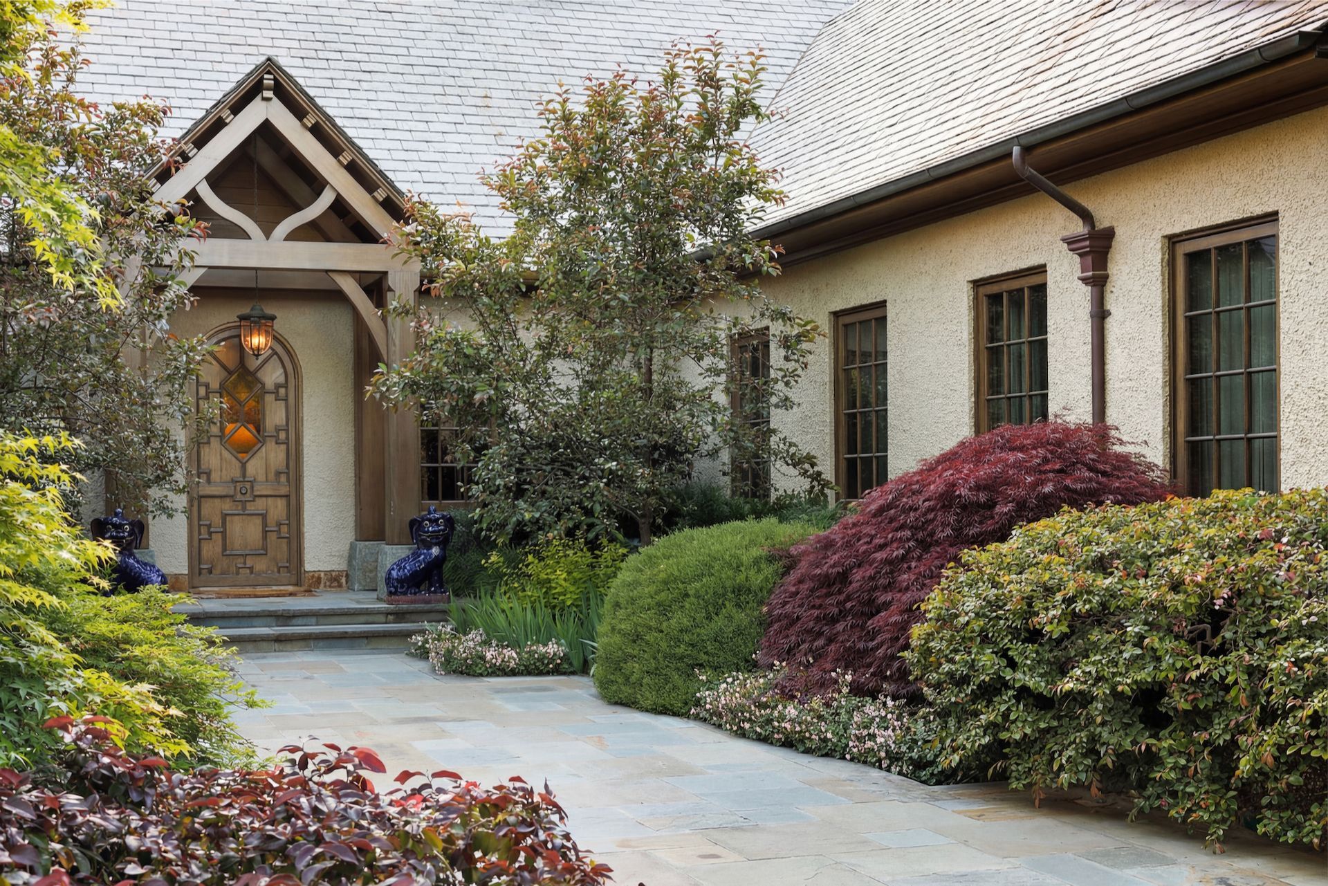 Stone pathway leads to a beige home with dark wood door and lush landscaping.