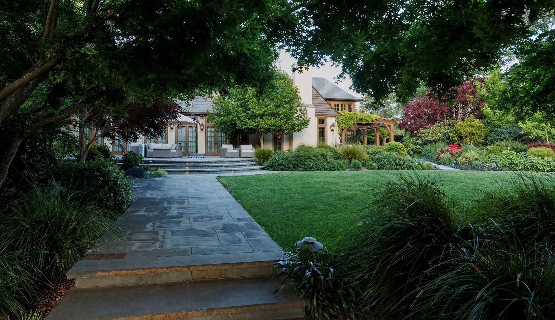 Stone path leads to a house with a large lawn and garden, surrounded by trees.