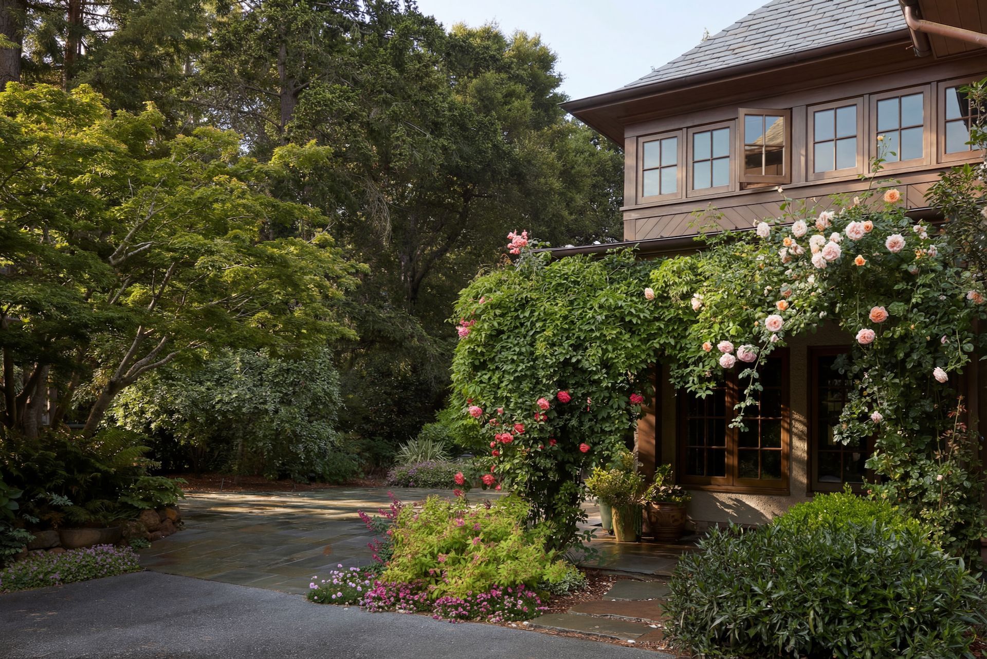 A house with a rose-covered archway; a stone path winds through a lush garden under a clear sky.