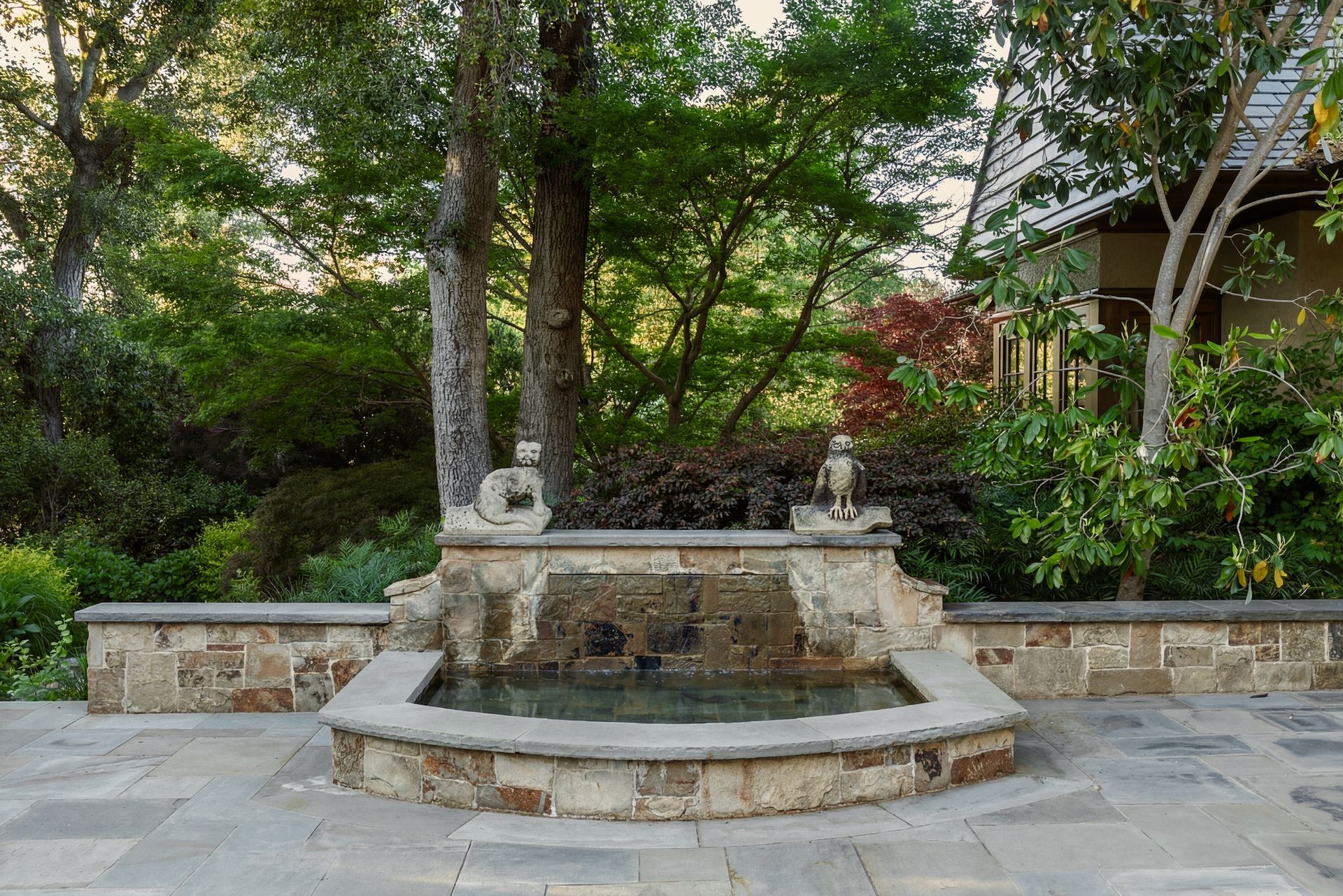 Stone fountain with statues in a garden setting, surrounded by trees and a building.