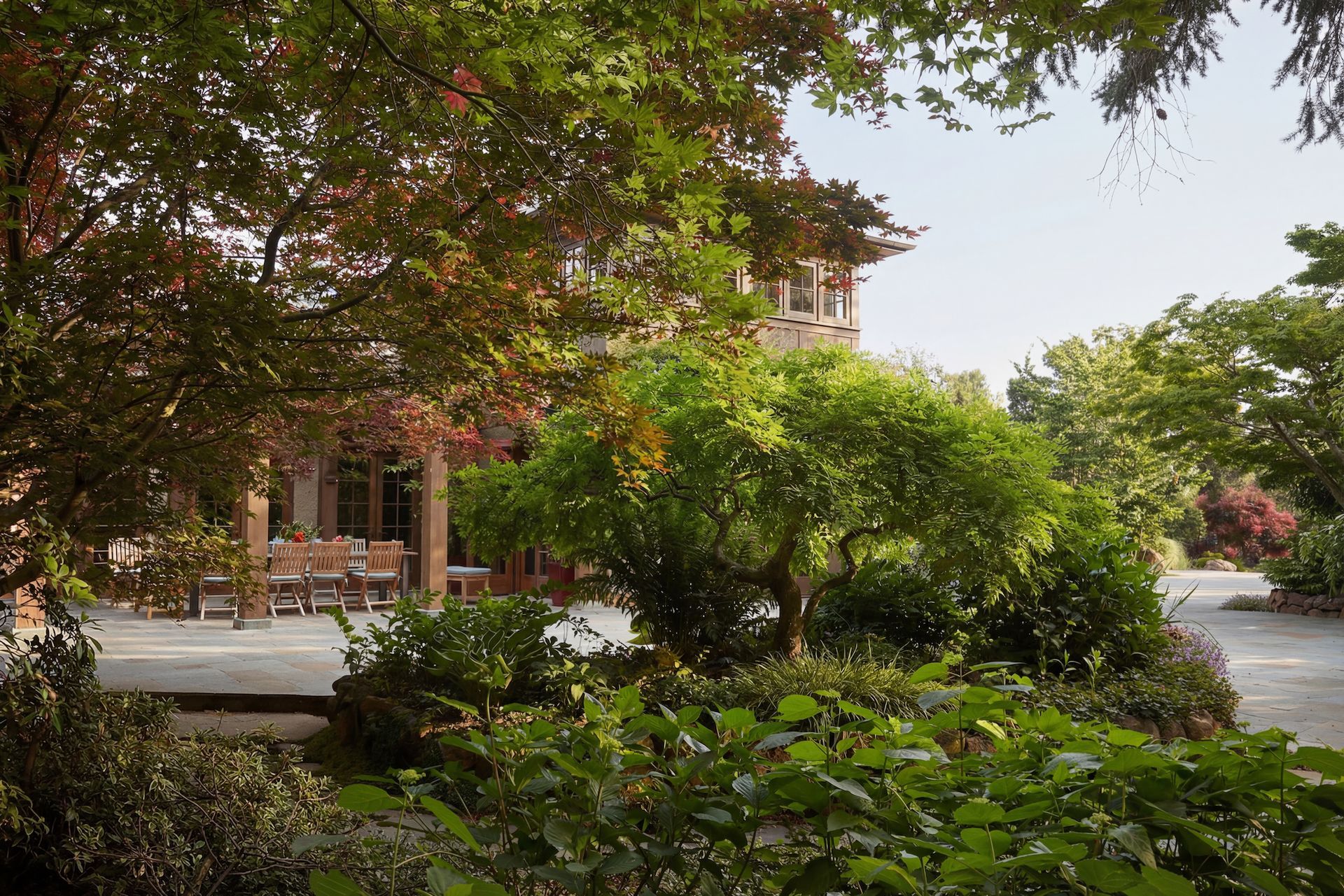 Lush garden with a house in the background. Green trees and plants frame a stone patio with outdoor seating.