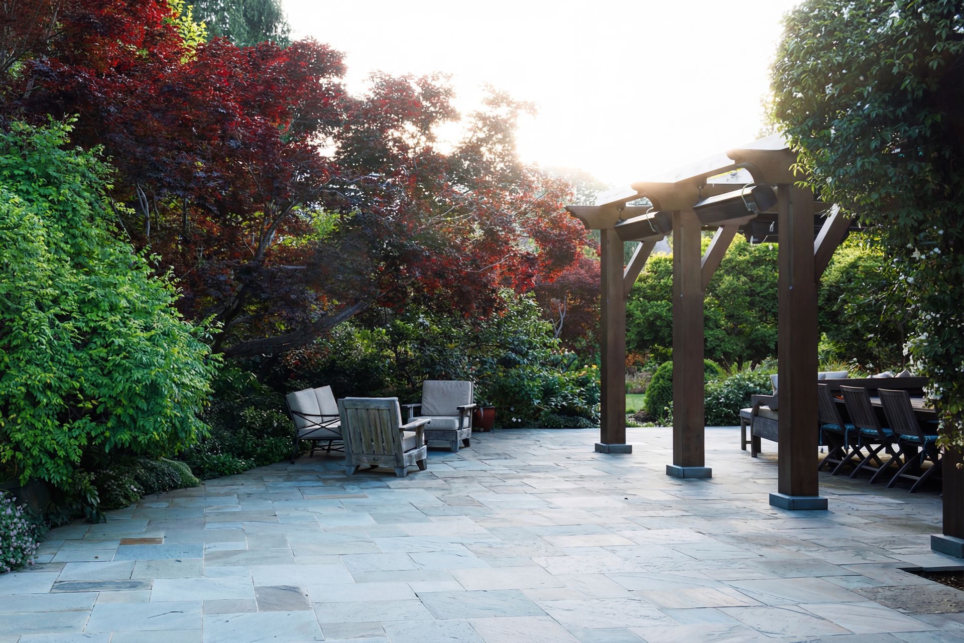 Patio with seating, pergola, and lush greenery in a garden. Sunlight shines through the trees.