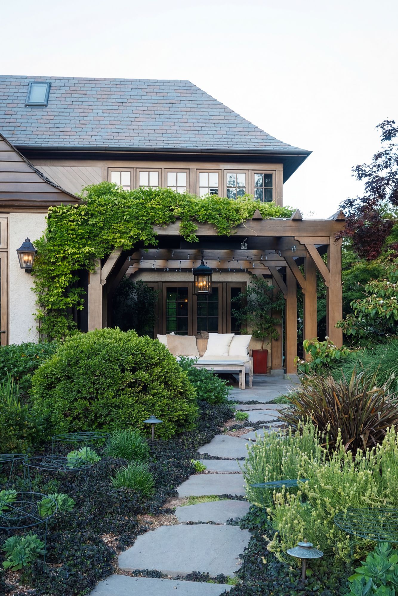Stone path leads to a wooden pergola with seating, covered in green vines, next to a house with a slate roof.