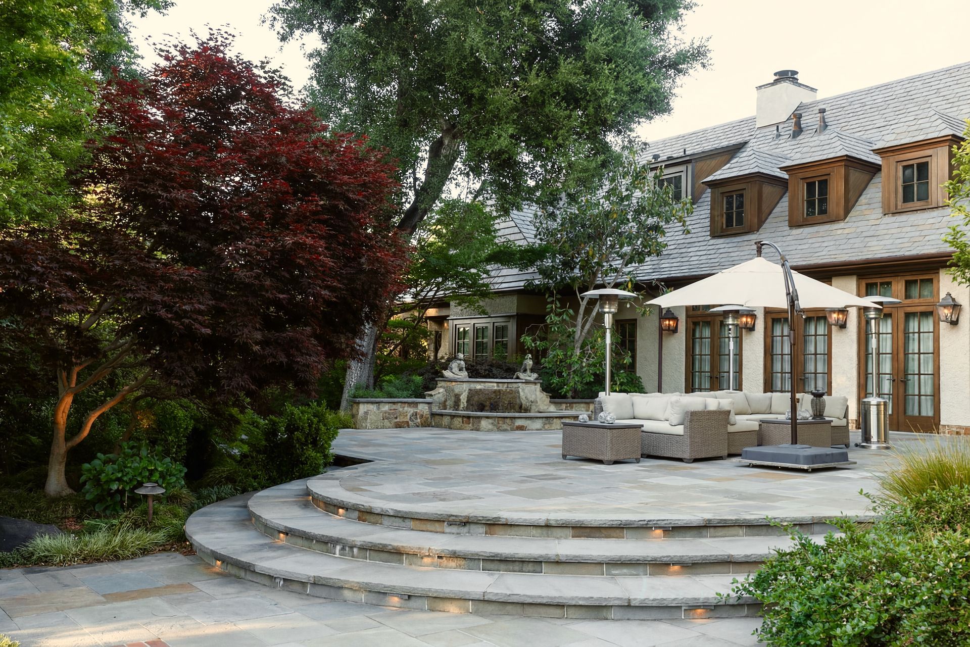 Stone patio with tiered steps, outdoor furniture, and fountain against a large house with a tile roof.