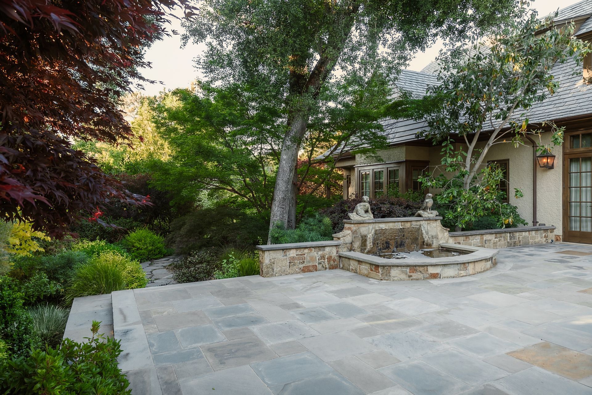 Stone patio with fountain, surrounded by lush greenery and a light-colored building.
