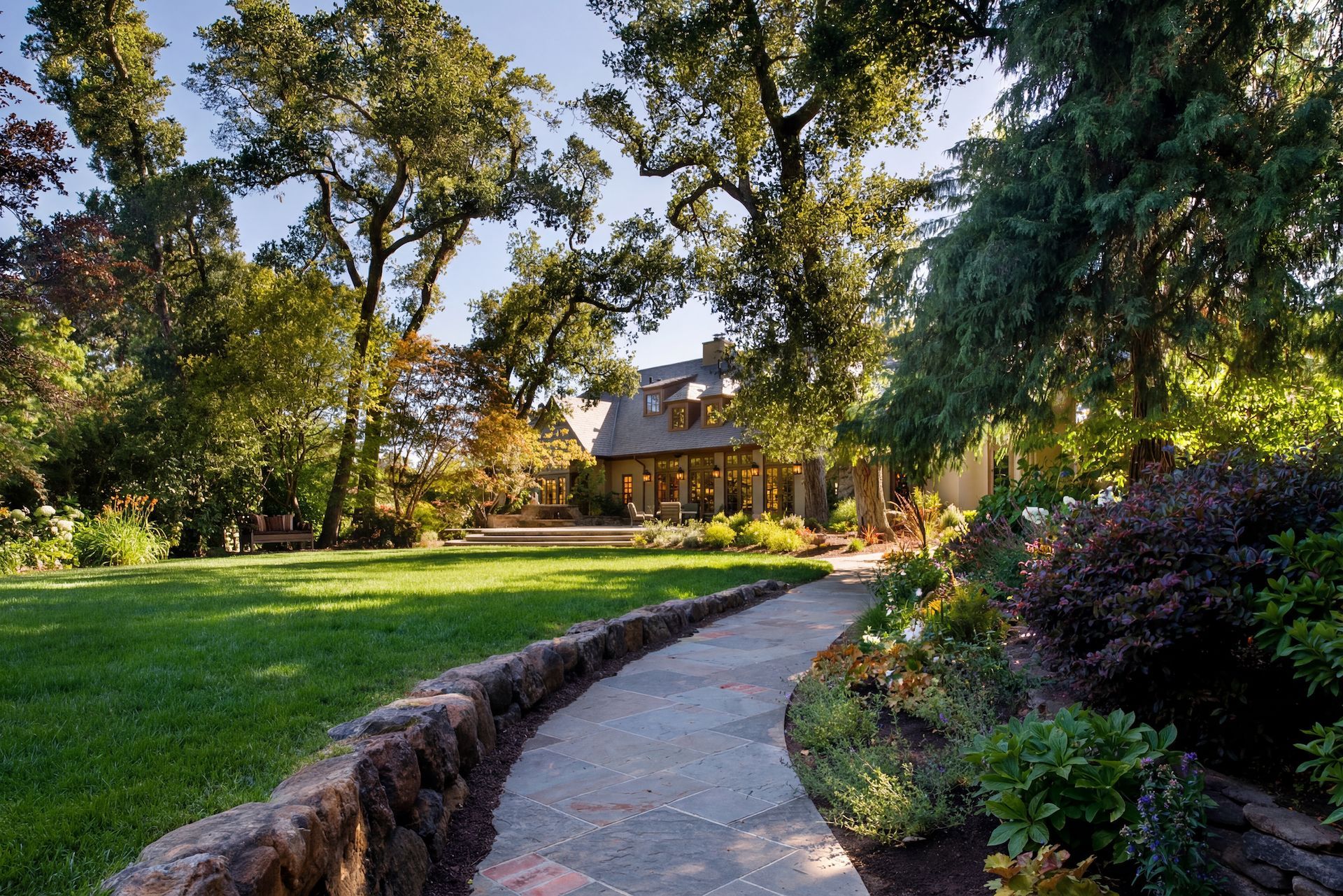 Stone path leads through a lush green garden towards a house partially obscured by trees.