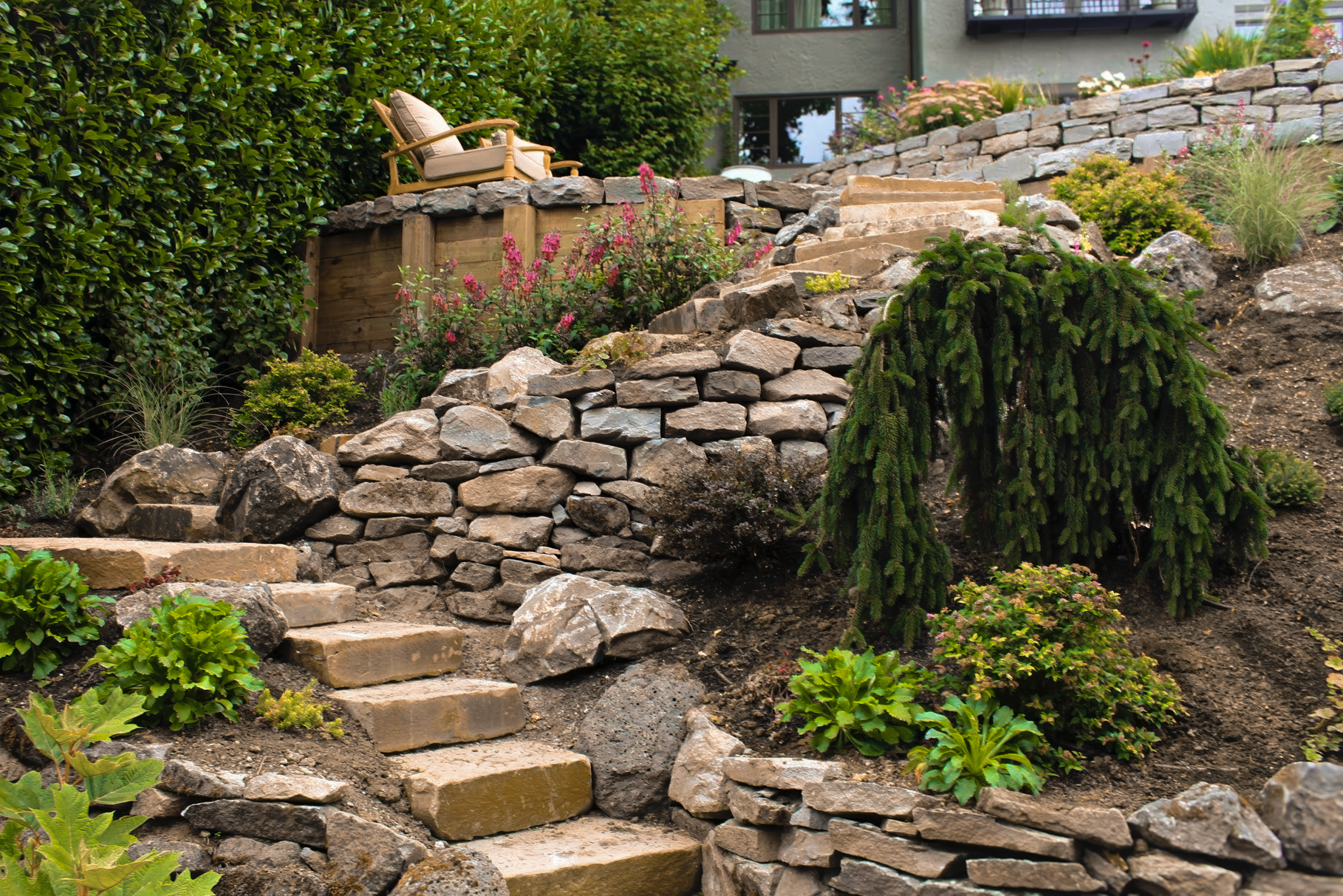 Stone steps and retaining walls in a landscaped hillside garden, with plants and a seating area.