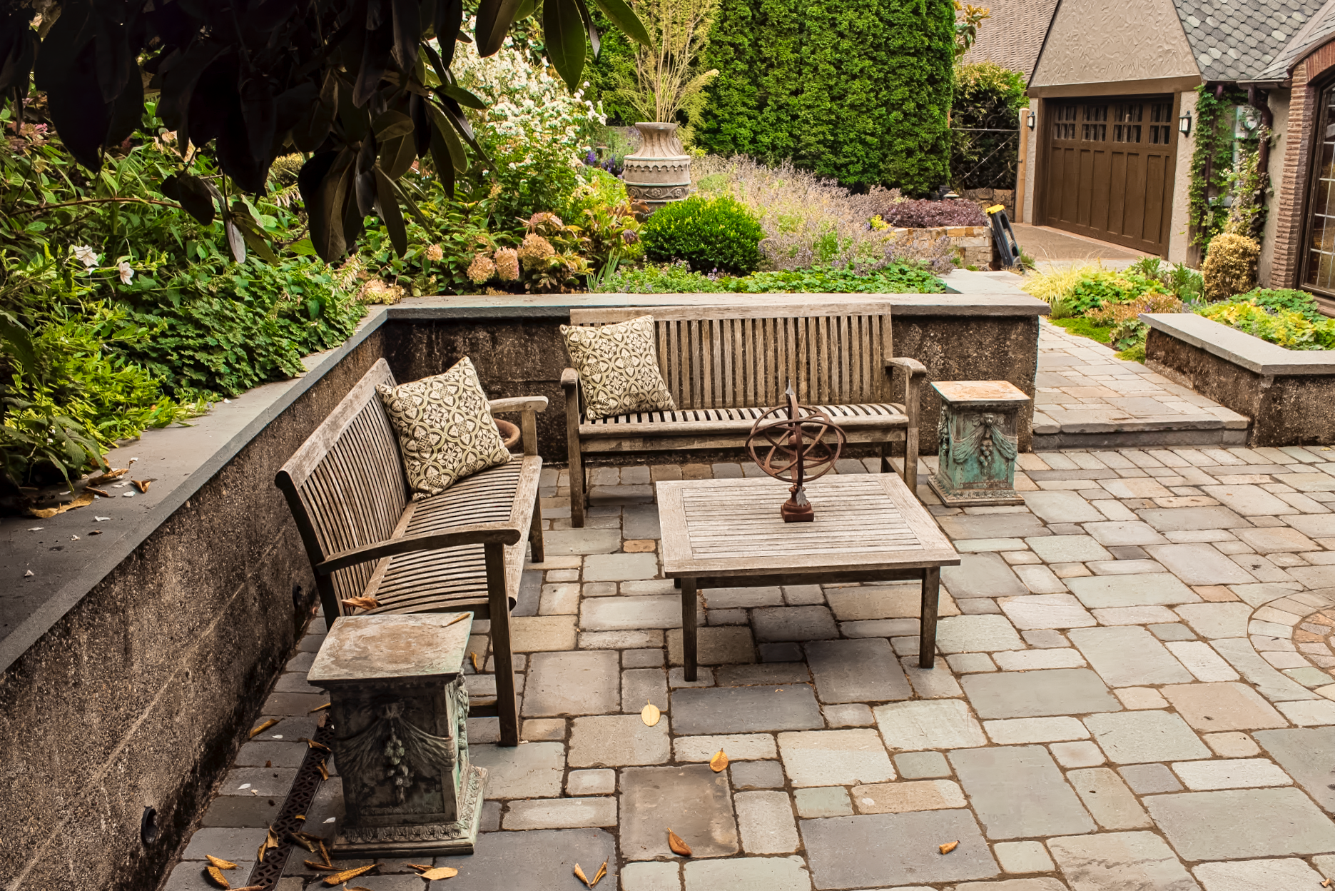Patio with wooden benches, table, and stone pavers surrounded by greenery and a garage.