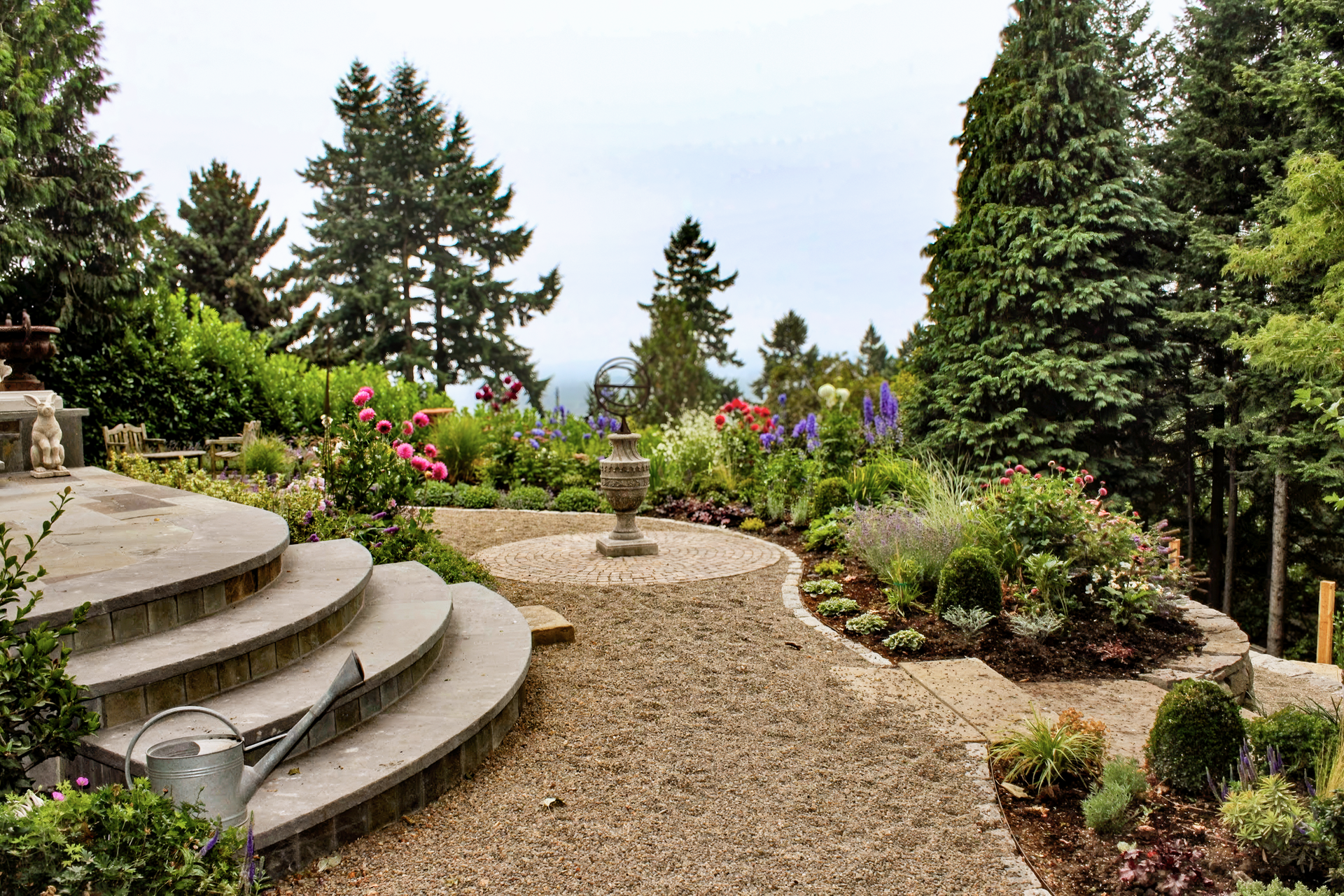 Stone steps lead to a garden with gravel paths, flowering plants, and trees under a cloudy sky.