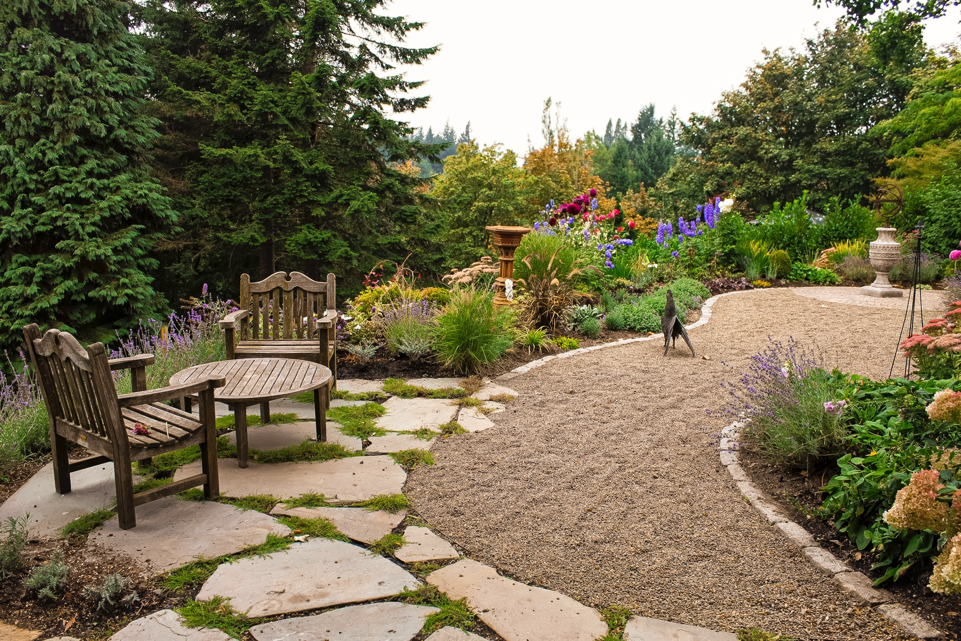 Wooden patio furniture in a lush garden with gravel path and colorful flowerbeds.