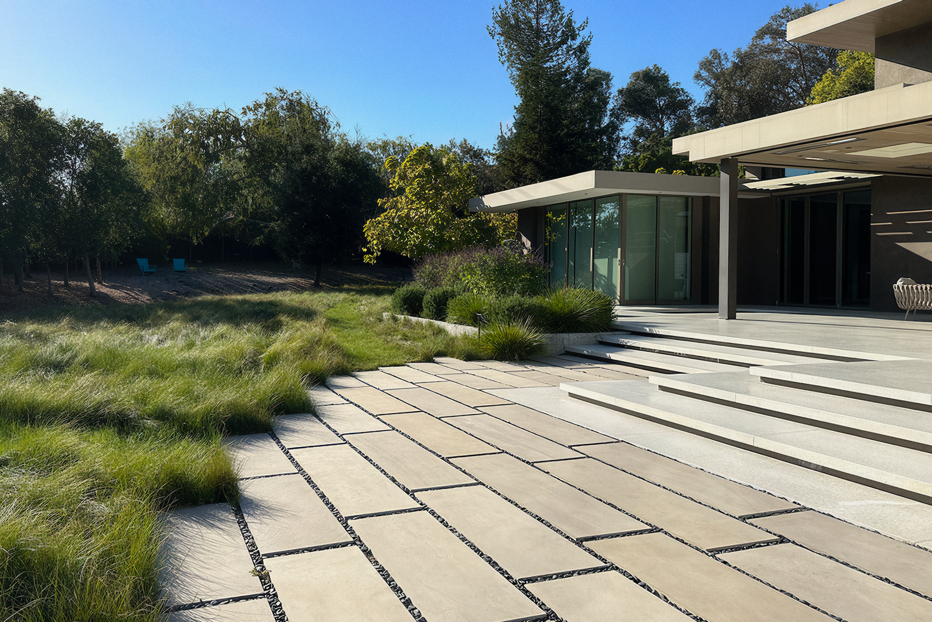 Stone patio and steps lead to a modern home with a glass enclosure; a grassy yard and trees in the background.