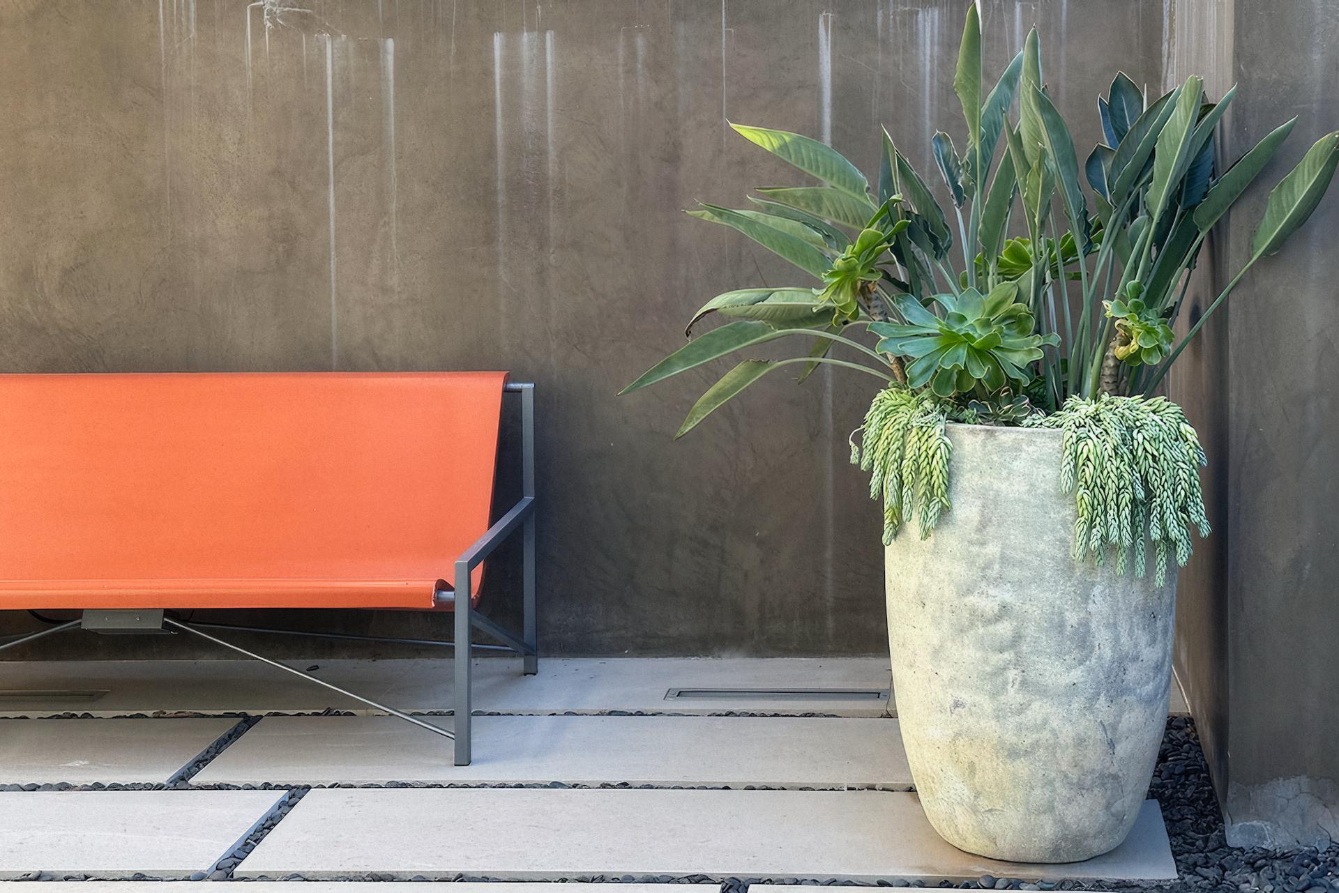 Orange bench, large stone planter with plants, gray concrete wall, outdoor setting.