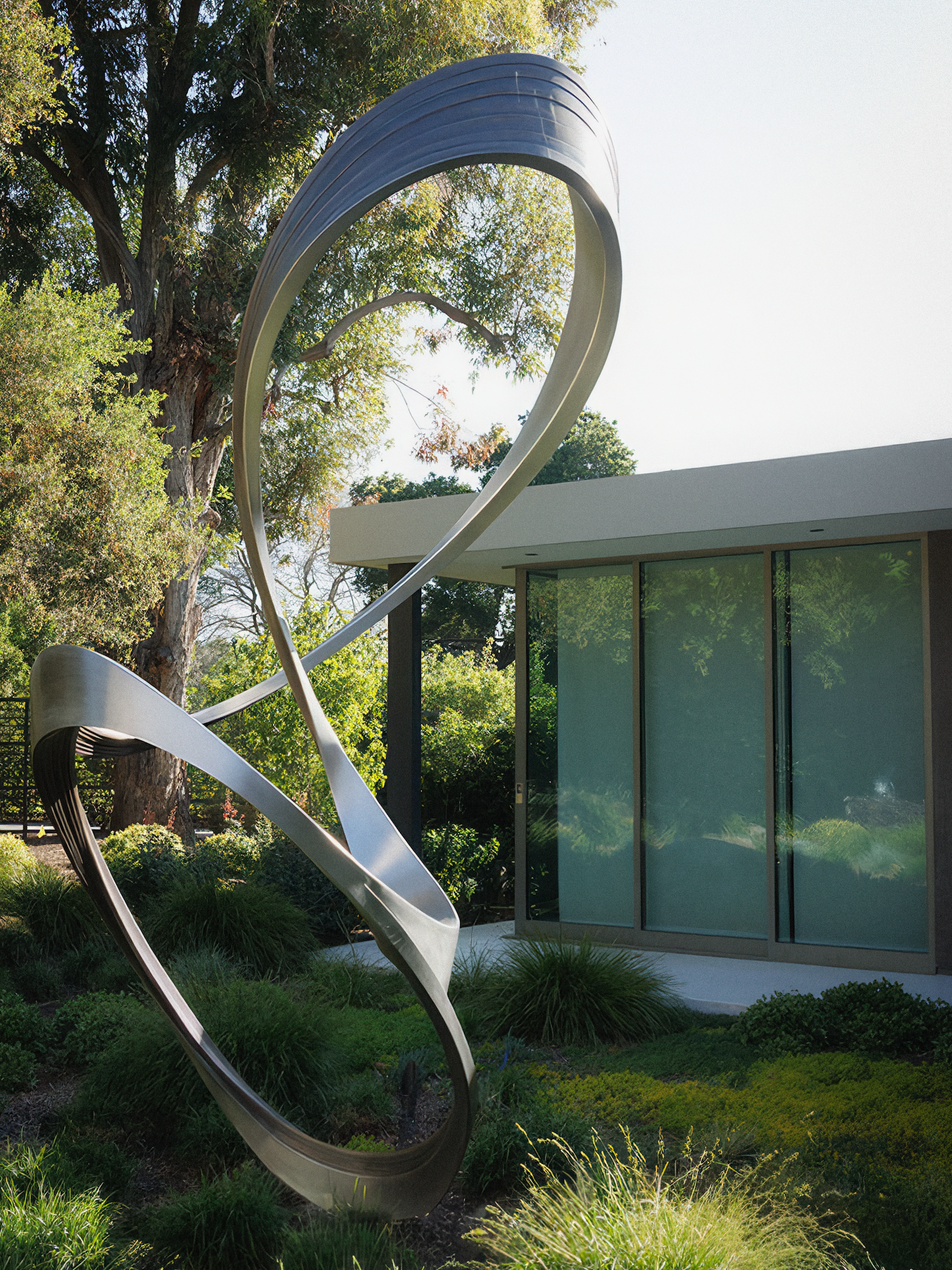 Abstract metal sculpture in a garden with a modern building and greenery in the background.