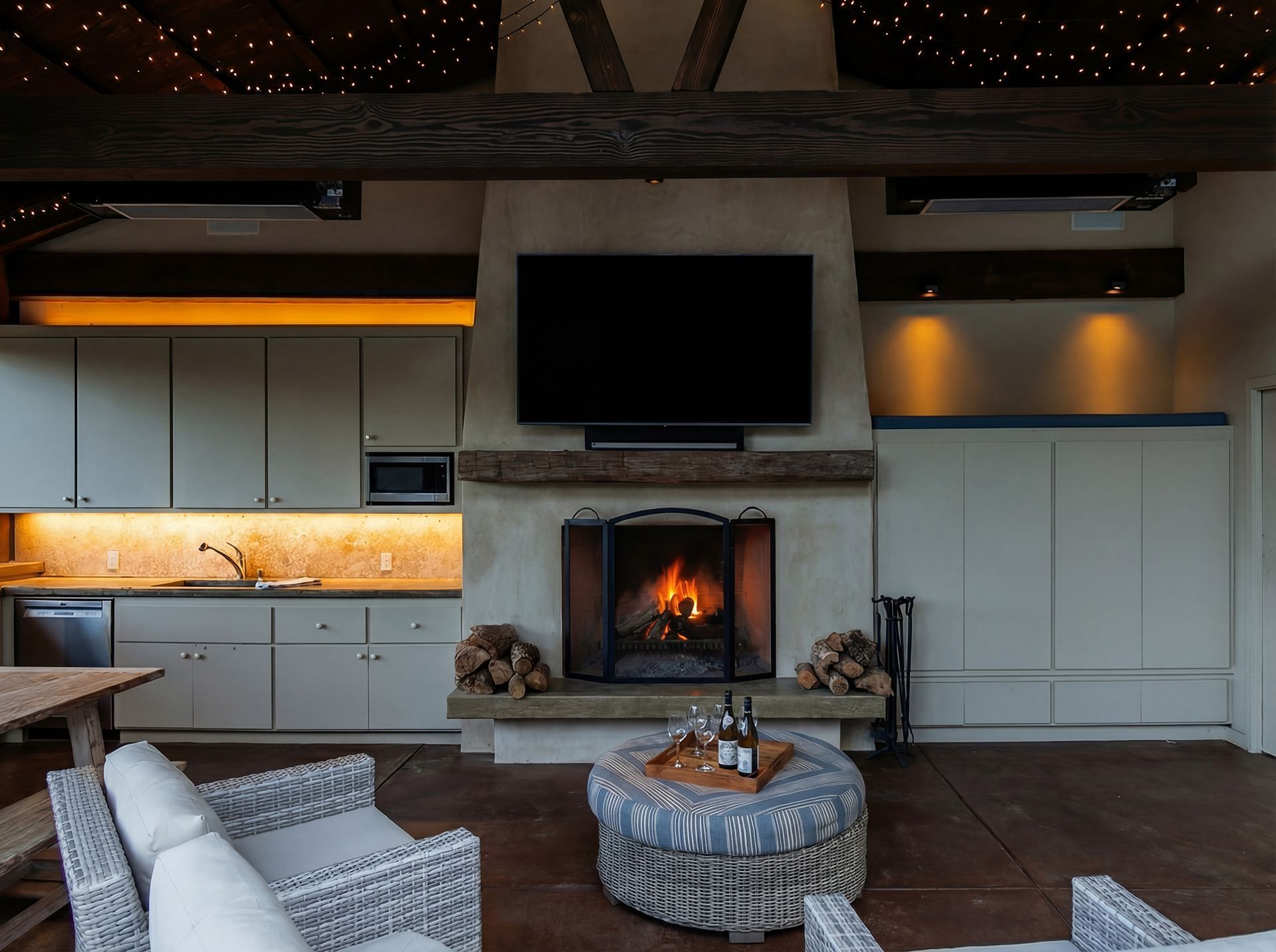 Cozy outdoor living area with fireplace, TV, kitchen, and seating. Fire lit in fireplace, string lights overhead.