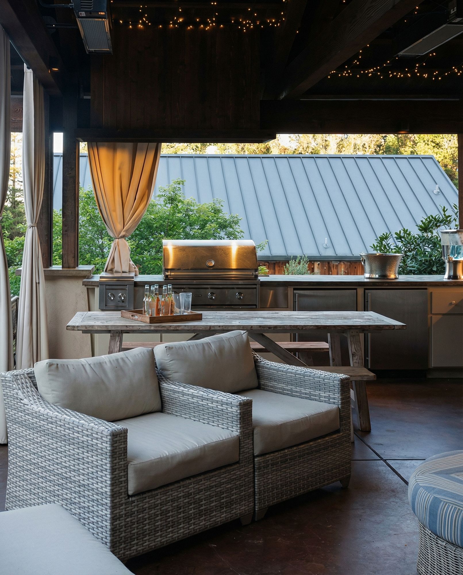 Outdoor kitchen area with a grill, table, wicker chairs, and neutral-colored curtains.