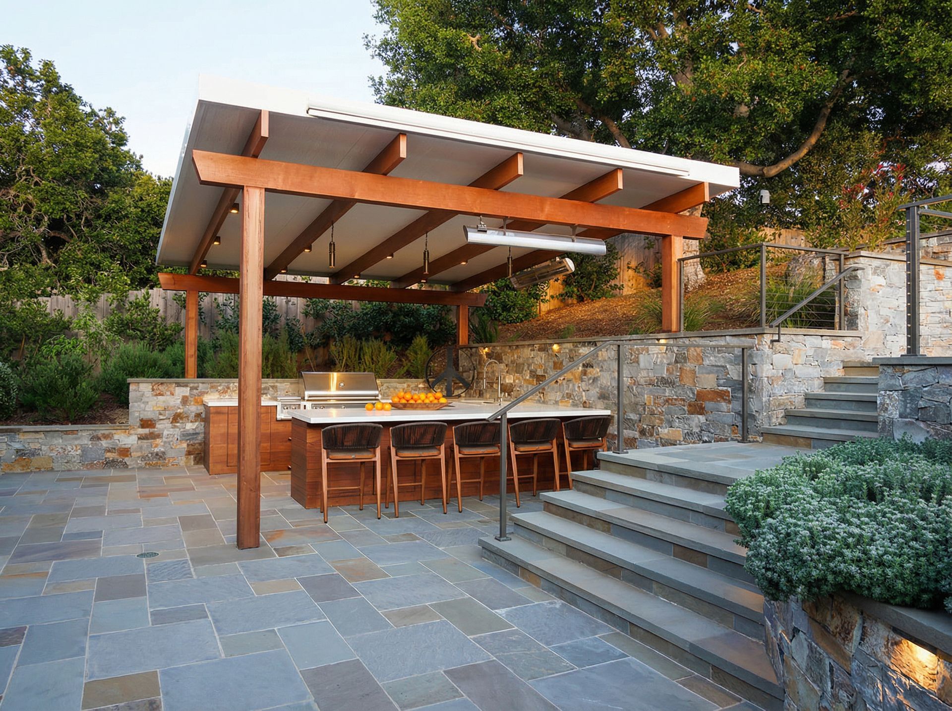 Outdoor kitchen under a wooden canopy with stone steps and patio.