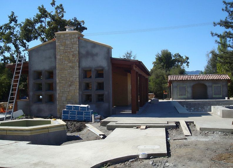 Construction site with two buildings and a chimney. Gray concrete walls, tan stone accents, blue tiles.