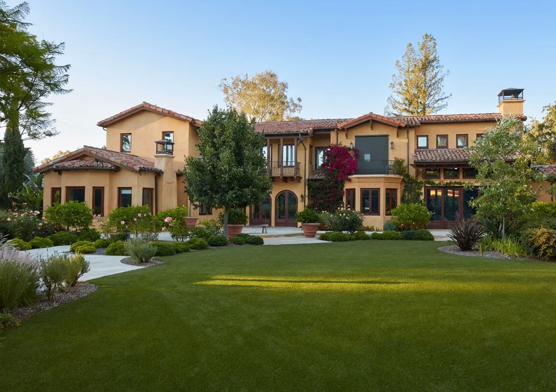 Large, beige-colored villa with terracotta roof tiles, surrounded by a green lawn and manicured landscaping