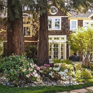 Brown shingled house exterior with flowering garden and large trees.