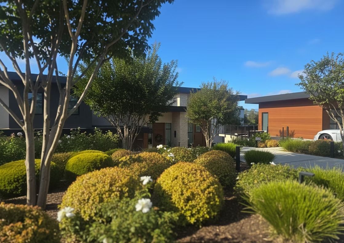 Well-manicured landscape with green and yellow bushes, trees, and modern buildings under a blue sky.