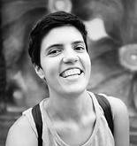 Person with short dark hair smiles widely outdoors, wearing a light tank top and backpack, floral background.