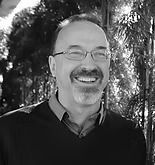 Man with glasses smiles; outdoors with trees in the background.