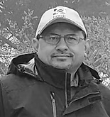 Man wearing glasses and a hat, smiling outdoors.