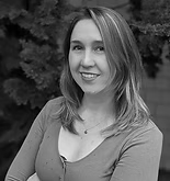 Woman smiling with arms crossed, wearing a low-cut shirt. Outdoors with foliage in background.