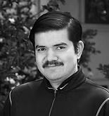 Man with mustache, wearing a dark jacket, smiles outdoors with foliage in the background.