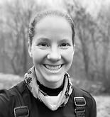 Woman smiling, wearing a patterned scarf and dark jacket outdoors, blurred background of trees.
