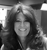 Woman with long, wavy hair smiles, looking at the camera. In a cafe, with blurred background and a hanging light.