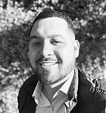 Man with dark hair and beard smiles, outdoors in front of leafy background.