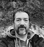 Man with a beard in a hooded jacket in front of a rocky background, looking at the camera.