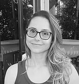 Woman with glasses smiling, leaning toward the camera, in front of a window.