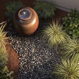 A round brown fountain in a garden bed of black pebbles and green plants.
