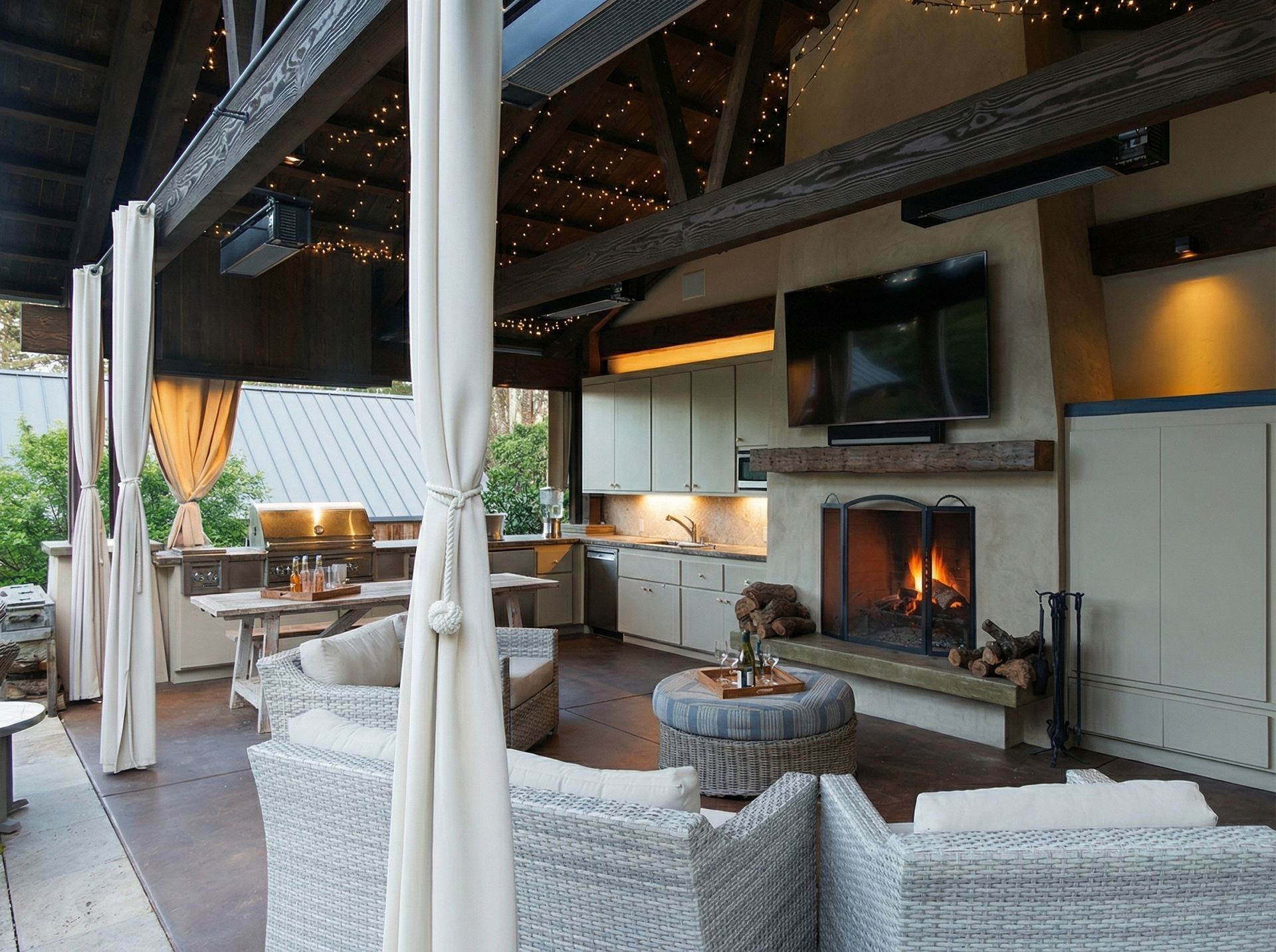 Outdoor living space with fireplace, kitchen, seating area, and curtains; lit with string lights.