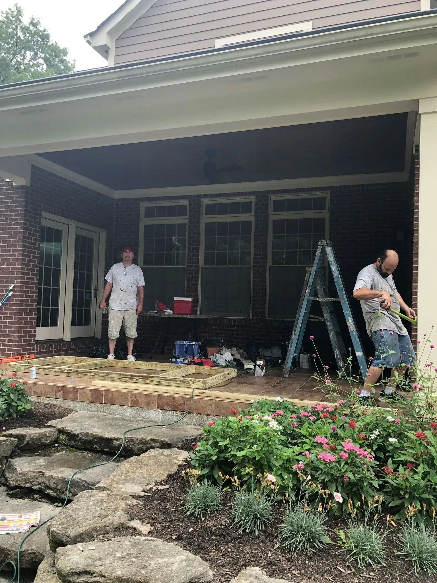 Two workers on a brick porch of a house; one stands, the other drills.
