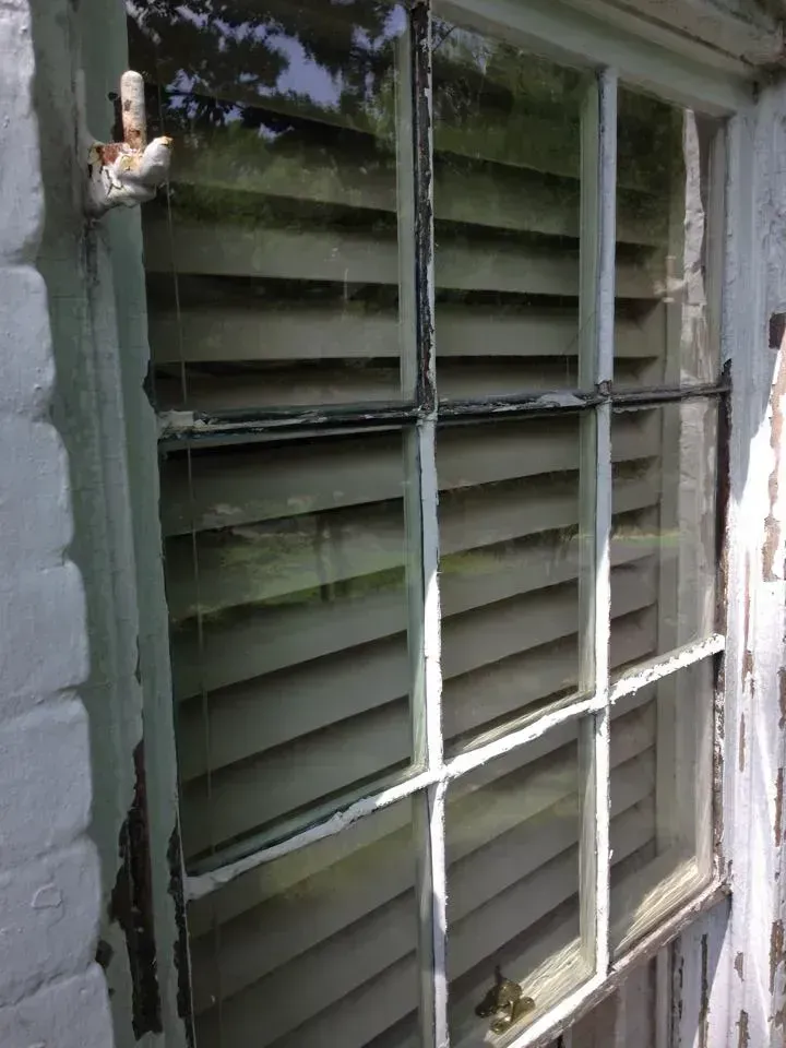 A weathered, white-framed window with interior shutters, a rusted hinge, and a blurred view of greenery.
