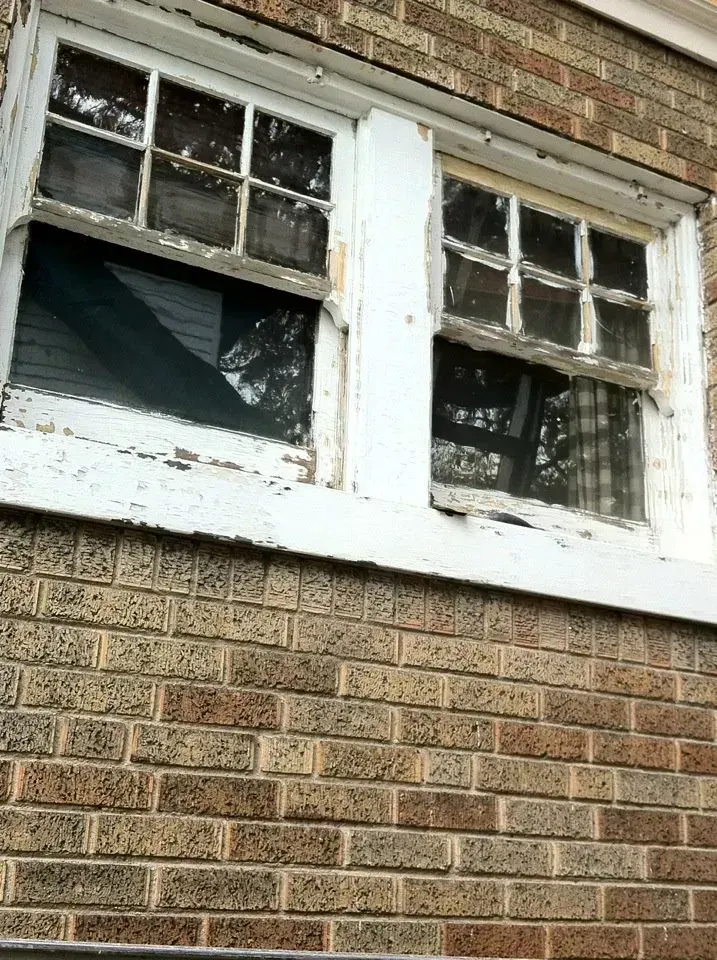 Two weathered white-framed windows set in a brown brick wall.