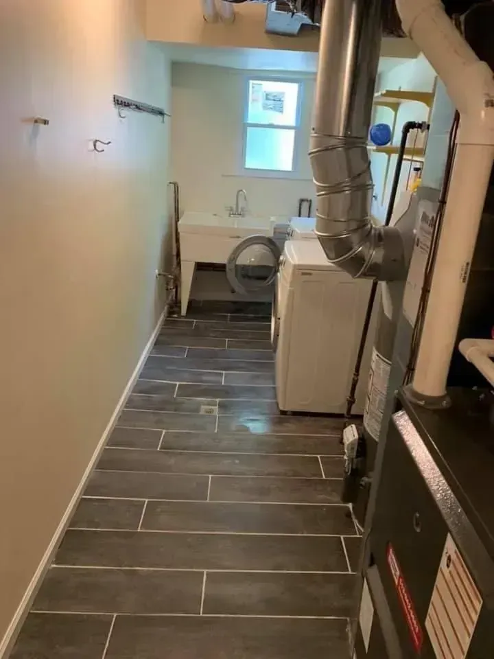 A narrow laundry room with dark tile floor, washer, sink, and furnace.