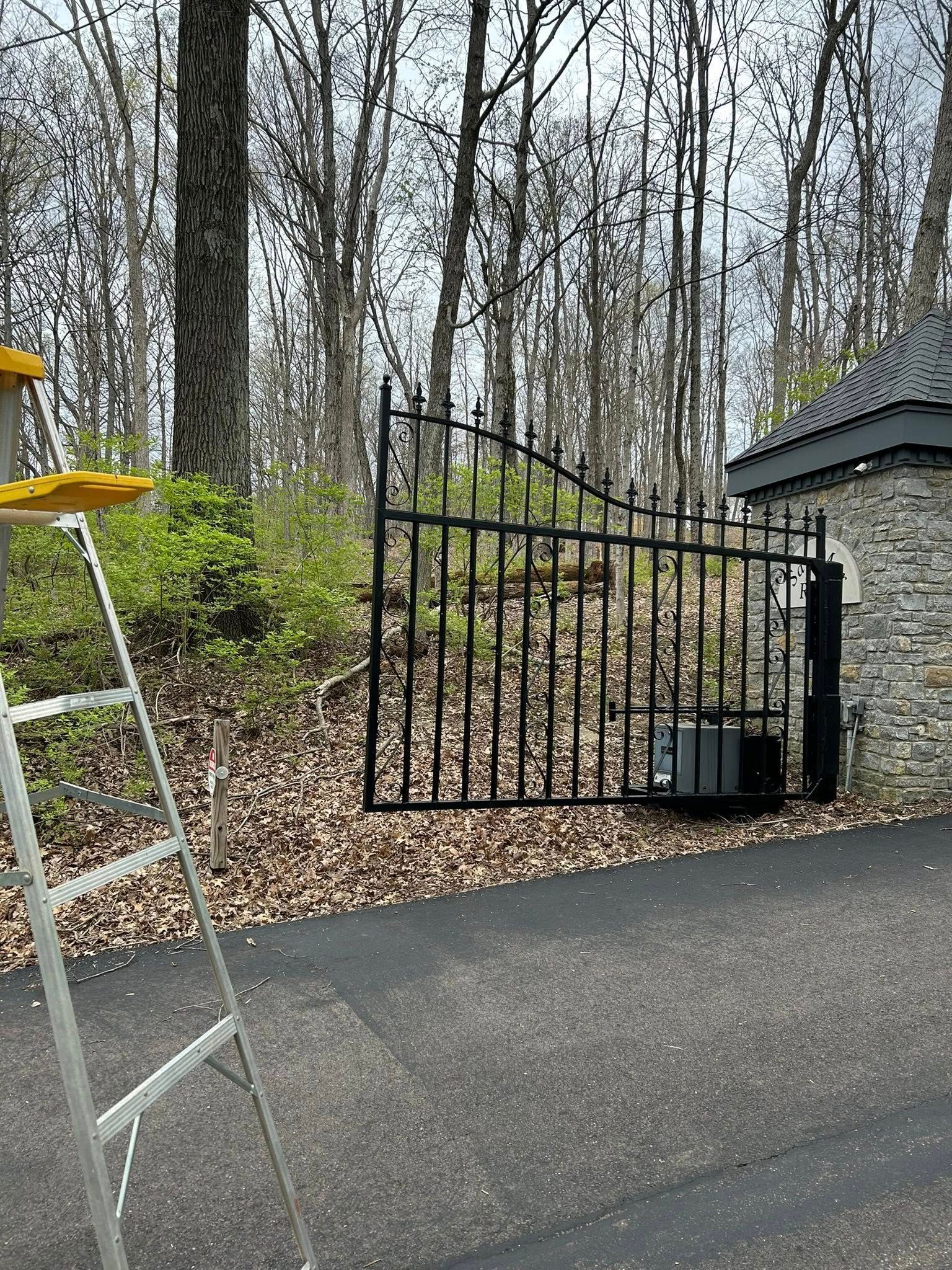 Black metal gate, set in stone pillars, closing a paved driveway in a wooded area.